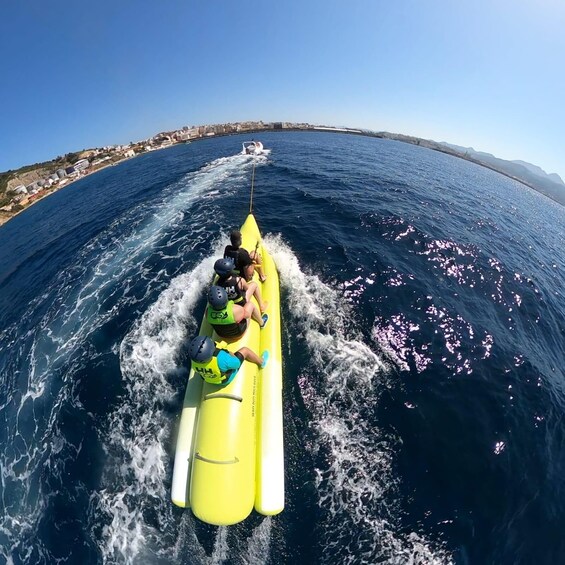 A yellow inflatable raft with three people wearing life jackets, being towed by a boat on a body of water with a coastline in the background. 3