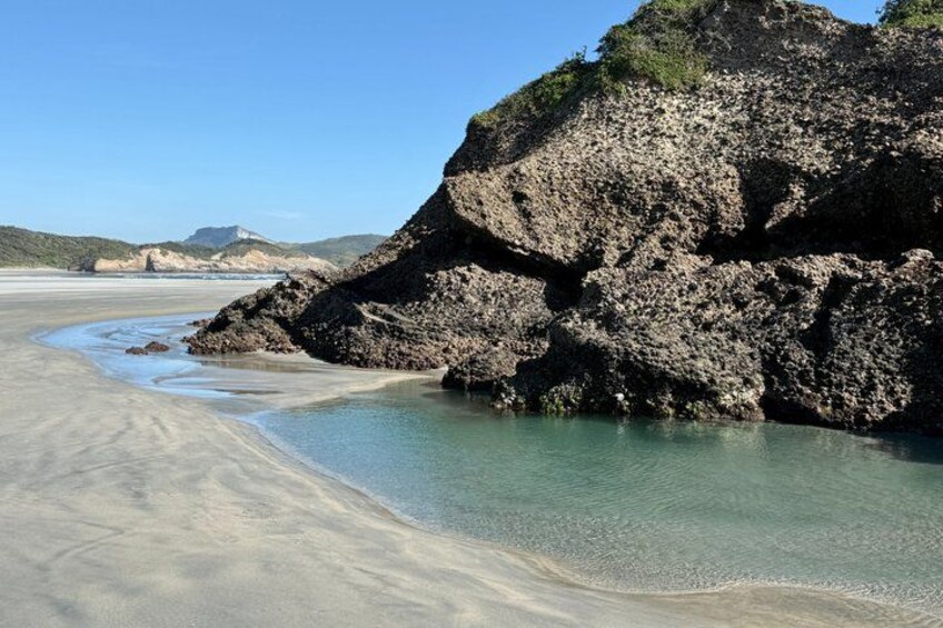 A coastal landscape with a rocky outcrop, clear water, and a sandy beach. 4