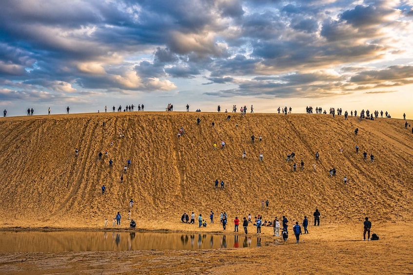 A large sand dune with people walking and standing on it, and a body of water at the base. 1