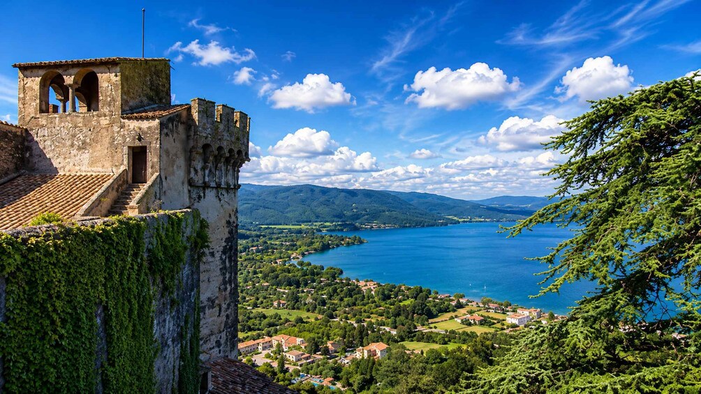 A historic stone tower with arches, a lush green landscape, a lake, and distant mountains under a clear blue sky. 2