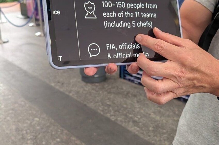 A person holding a tablet displaying text about a team event with 100-150 people from each of the 11 teams, including 5 chefs, and mentions FIA, officials, and official media. 4