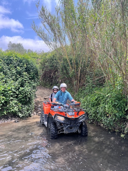 Two people on an ATV in a narrow water crossing surrounded by dense vegetation. 2