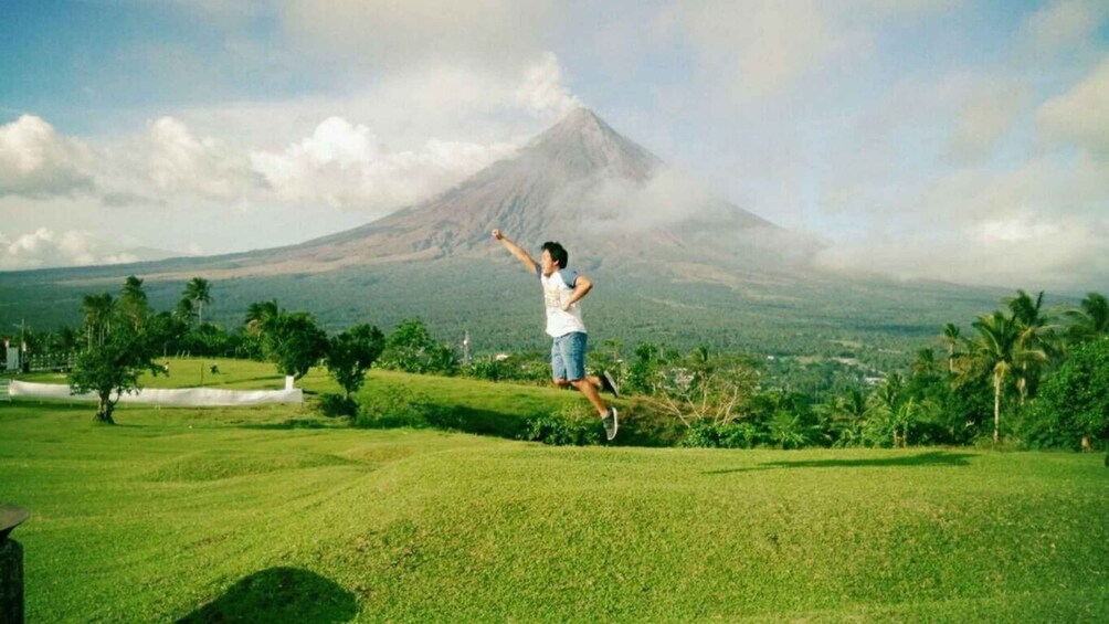 A person jumping on a grassy field with a volcano in the background. 3