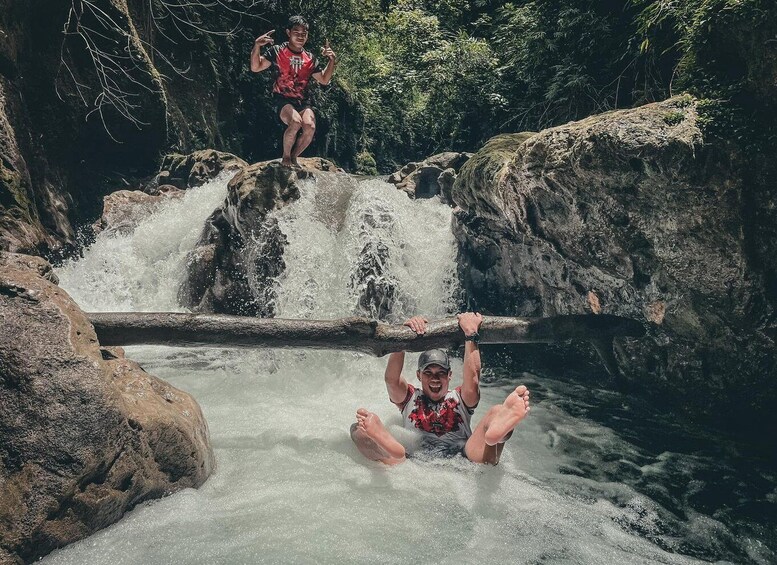 A waterfall with two people; one is standing on a rock above the water, and the other is swimming below. 1