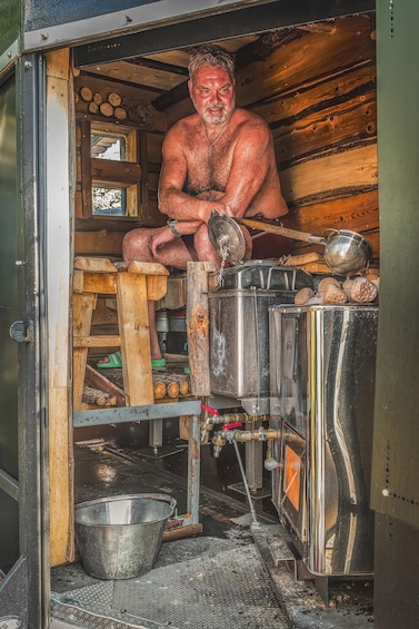 A man in a rustic cabin with a wood stove, metal pots, and a metal bowl. 1