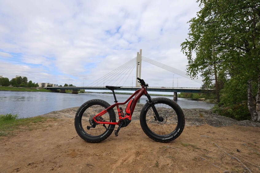 A red mountain bike parked on a sandy shore with a river and a bridge in the background. 5
