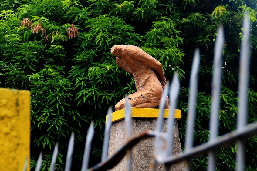 A bronze statue of a seated figure on a pedestal, surrounded by lush green foliage. 1