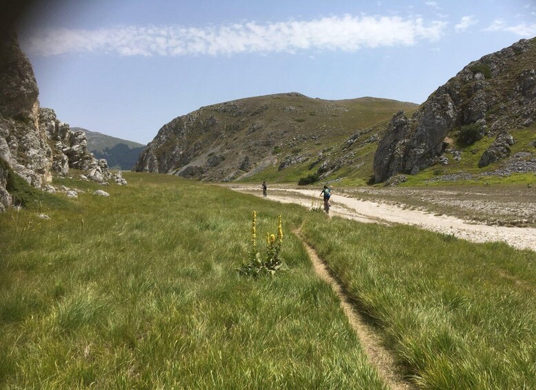 A hiker on a dirt trail surrounded by grassy hills and rocky cliffs. 2
