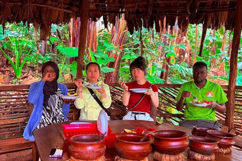 Four people sitting at a wooden table with plates of food, surrounded by large clay pots and a thatched roof structure. 3