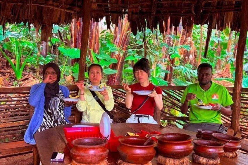 Four people sitting at a wooden table with plates of food, surrounded by large red bowls and a tropical garden backdrop. 3