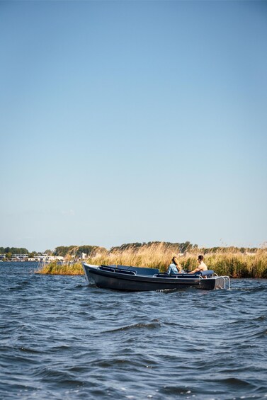 A small boat on calm water with two people, surrounded by tall grasses and a clear sky. 1