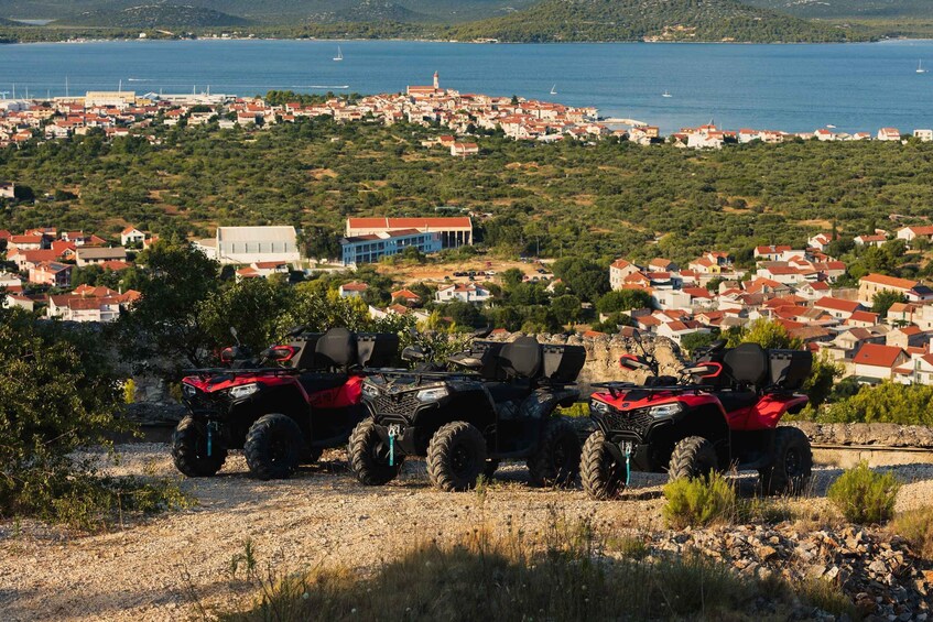 A coastal town with red-roofed houses, a body of water, and three red all-terrain vehicles parked on a gravel path. 3