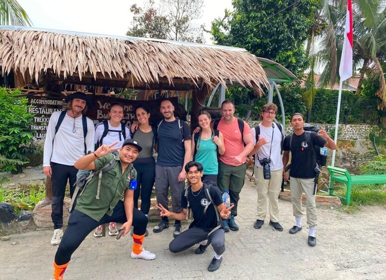 A group of people posing in front of a rustic hut with a thatched roof, surrounded by greenery and a stone wall. 2