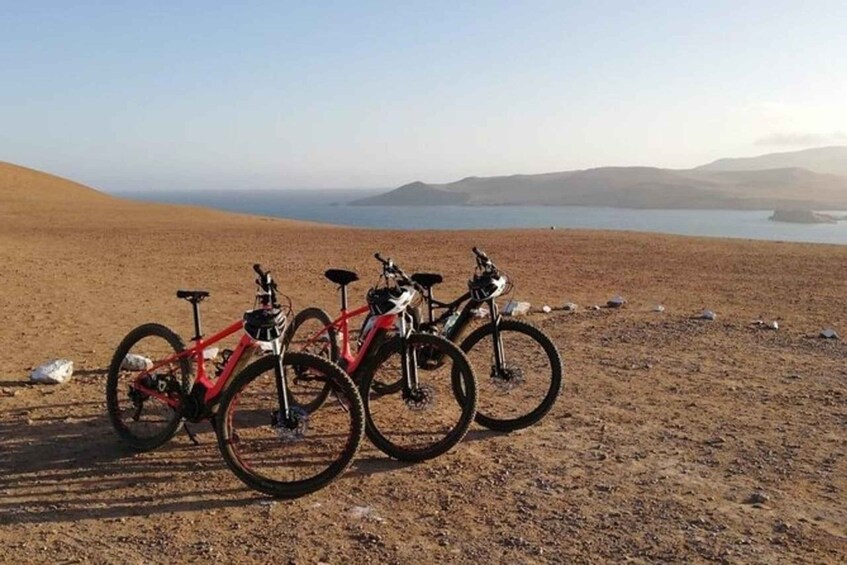 Two bicycles parked on a sandy beach with a body of water and hills in the background. 2