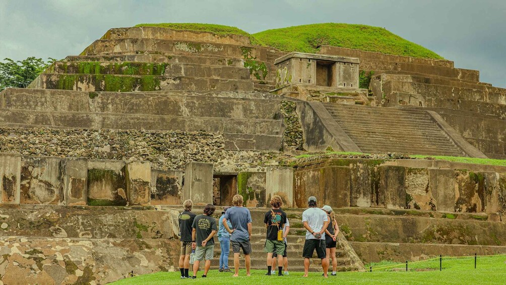 A group of people standing in front of ancient ruins with stepped structures and a small pyramid-like building. 5