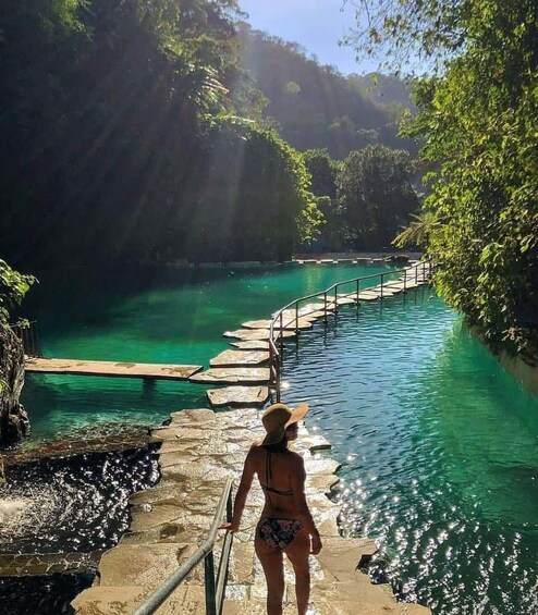 A woman in a bikini walking on a stone pathway beside a clear blue pool surrounded by lush greenery. 1