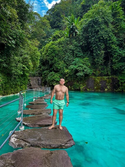 A man standing on a rock platform in a turquoise pool surrounded by lush greenery. 2
