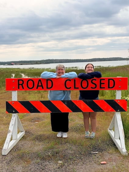 Two people standing behind a road closure sign with a lake and forest in the background. 5
