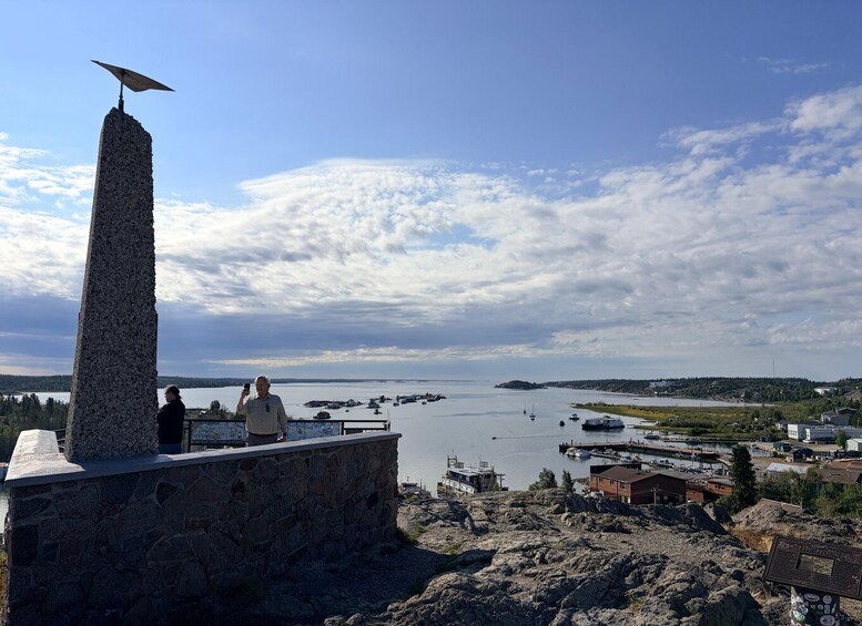 A rocky outcrop with a tall monument topped with an umbrella, overlooking a calm body of water with boats and a distant shoreline. 1