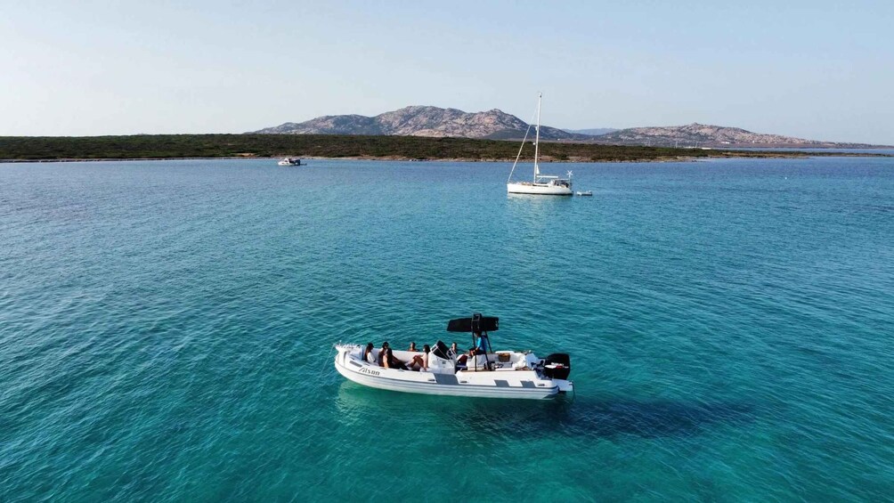 A boat with people on a turquoise sea, a sailboat in the distance, and mountains in the background. 1