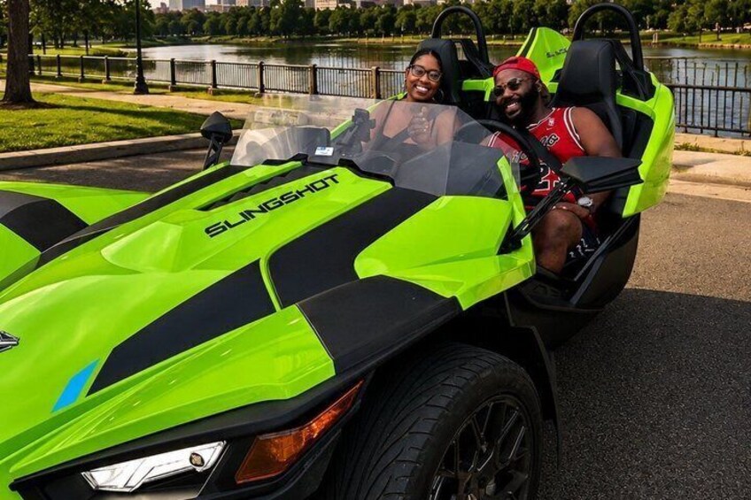 A bright green and black vehicle with the text 'SLINGSHOT' on the hood, parked on a paved area near a body of water. 3