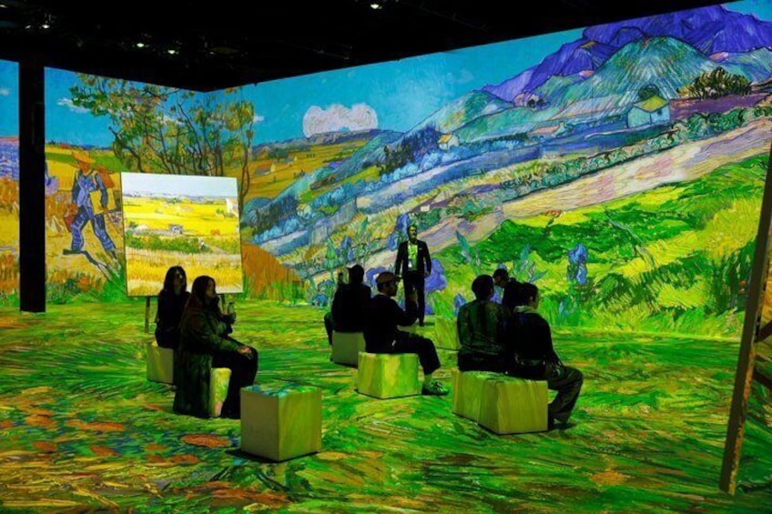 A group of people seated on stools in a room with a large painting of a landscape. 2