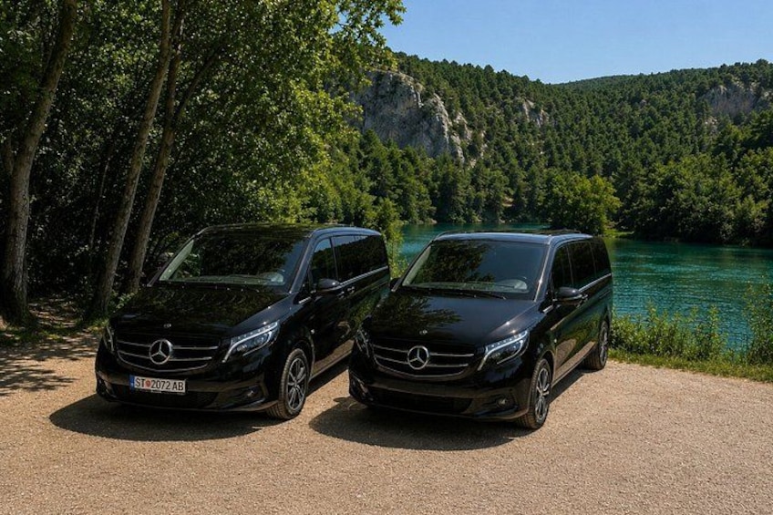 Two black Mercedes-Benz vans parked on a gravel path beside a lake with trees and cliffs in the background. 1
