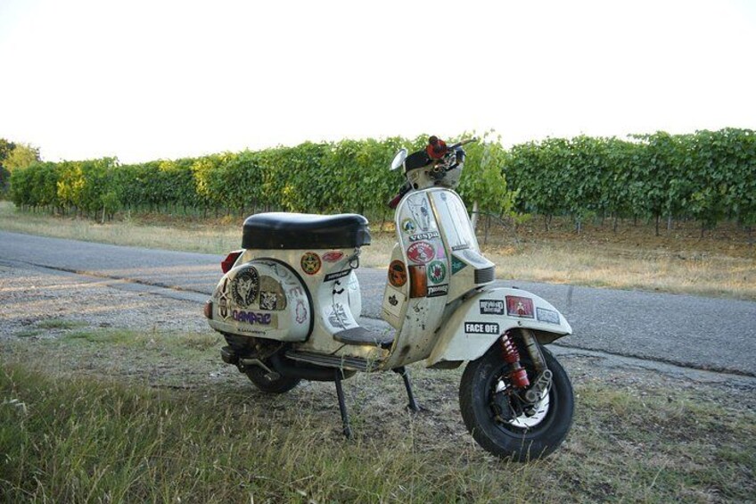 A white scooter with various stickers parked on a grassy roadside. 3