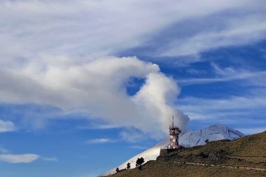 A mountain with a tower emitting smoke, a clear sky, and a snow-capped peak. 1