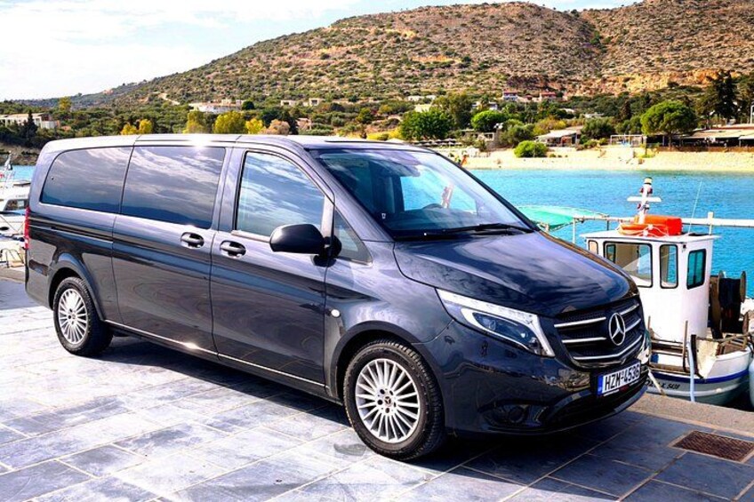 A dark-colored Mercedes van parked near a body of water with boats and a hilly landscape in the background. 3