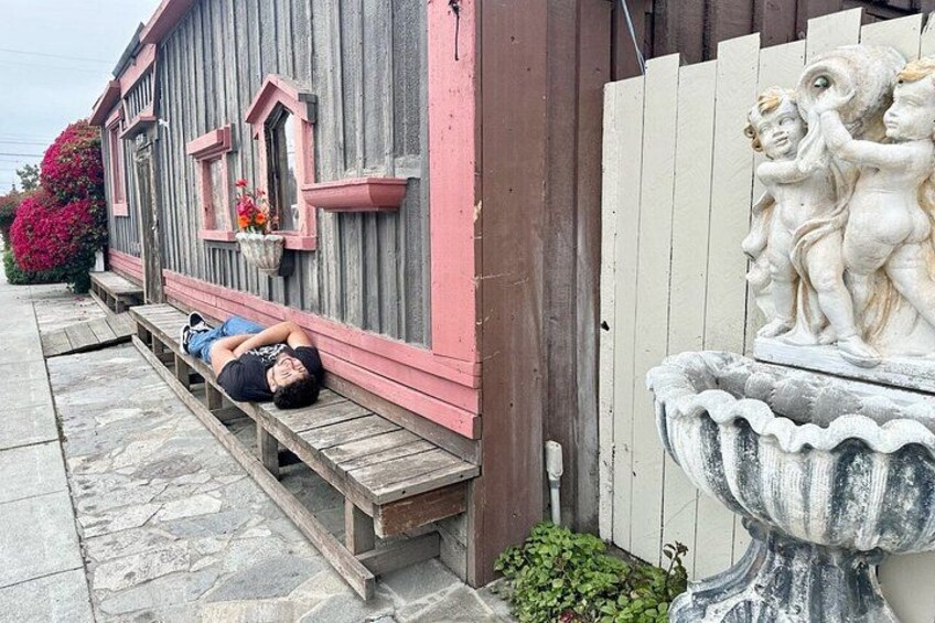 A person lying on a wooden bench outside a building with a pink and gray facade, a stone pathway, and a white fence with a statue of cherubs. 3