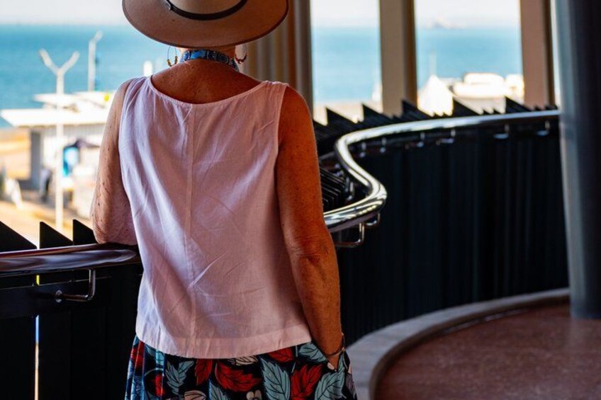 A person wearing a hat and a sleeveless top, standing near a railing with a view of the ocean. 1