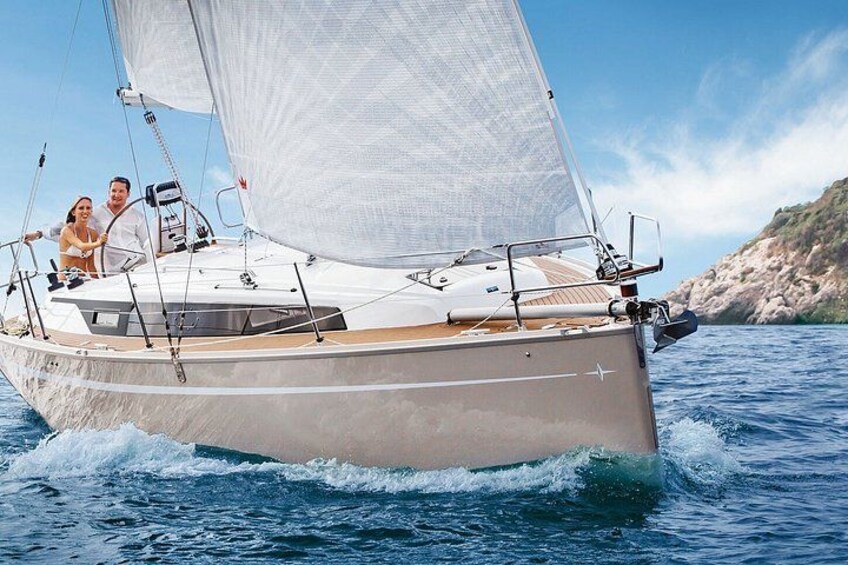A sailboat with a white hull and a person steering, sailing on blue water with a rocky coastline in the background. 2