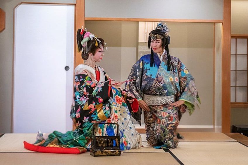 Two individuals in traditional Japanese attire, sitting on tatami mats with a red object and a patterned bag on the floor. 2