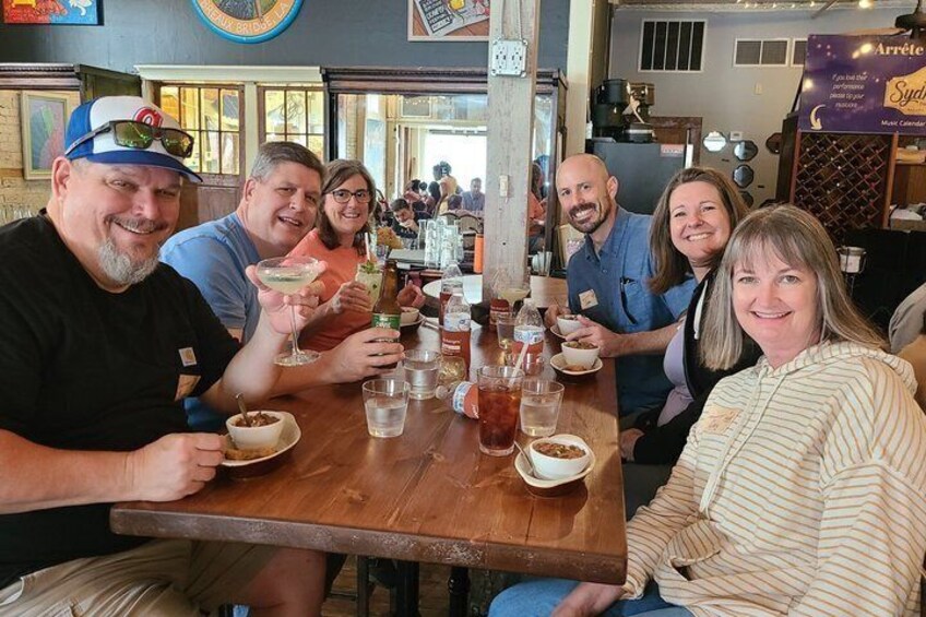 A group of people sitting at a wooden table with drinks and food, in a restaurant with a brick wall and a clock. 1