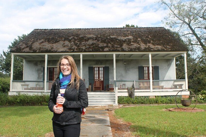 A woman standing in front of a historic house with a porch, a garden, and a large tree. 2