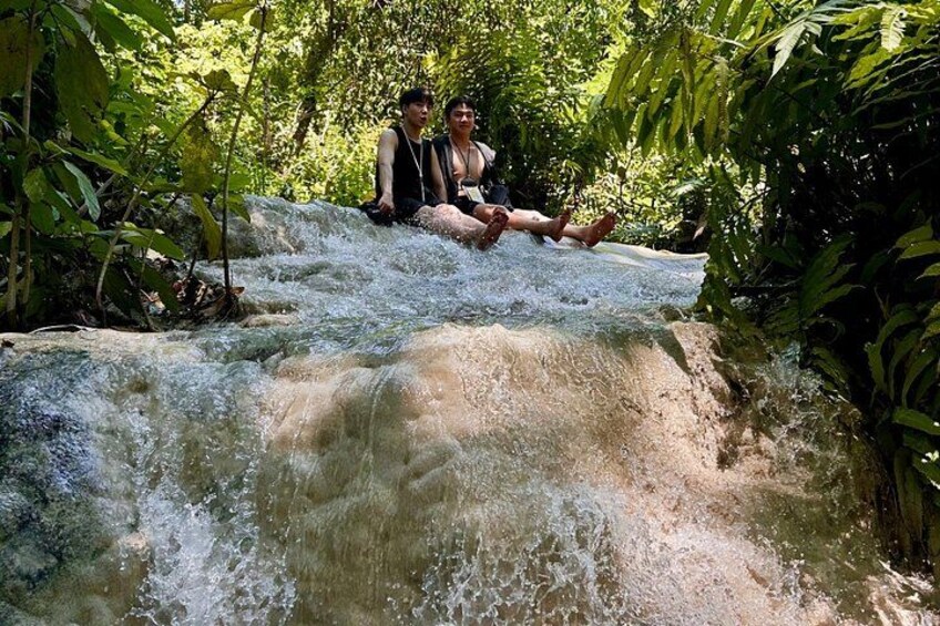 Two people sitting on a rock by a flowing waterfall surrounded by dense greenery. 2
