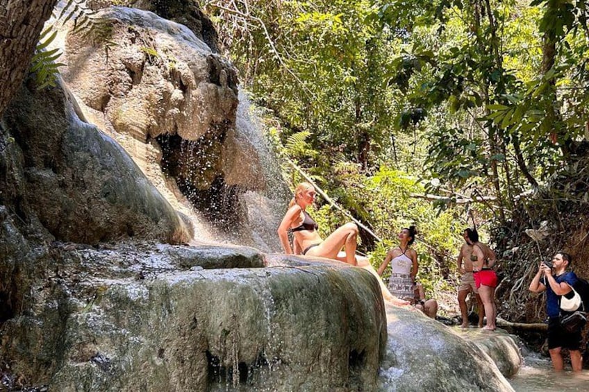A waterfall in a forested area with people sitting and standing around. 1