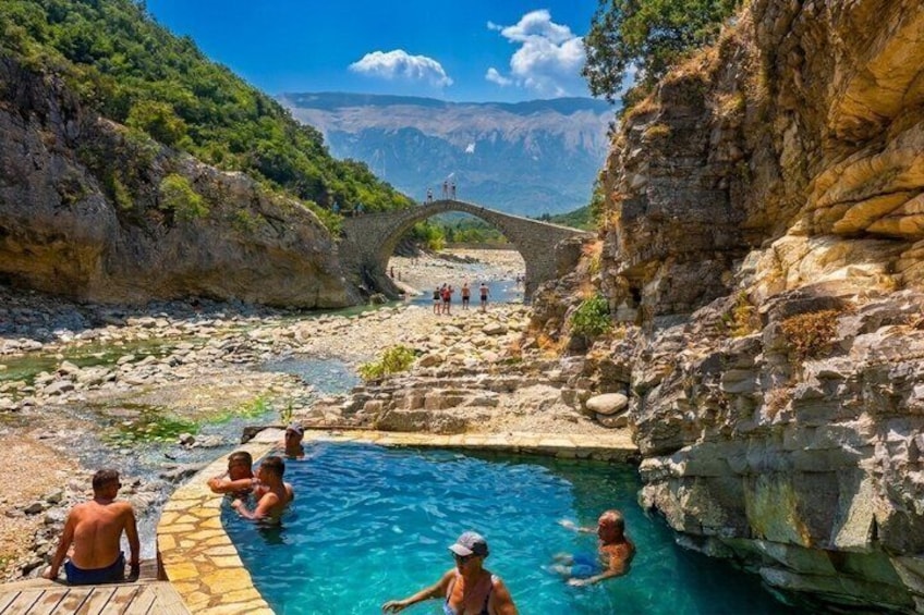 A natural swimming pool surrounded by rocky cliffs, a stone bridge, and a mountainous backdrop. 1