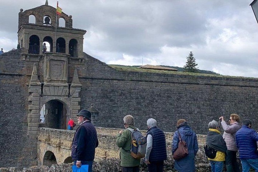 A stone wall with a bell tower, people standing and looking at it, and a cloudy sky. 1