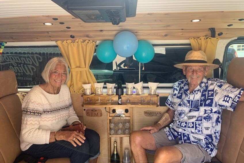 Two people seated in a vehicle interior with a wooden ceiling, a window with curtains, and a table with various items including a bottle and a box labeled 'SIMI'. 5