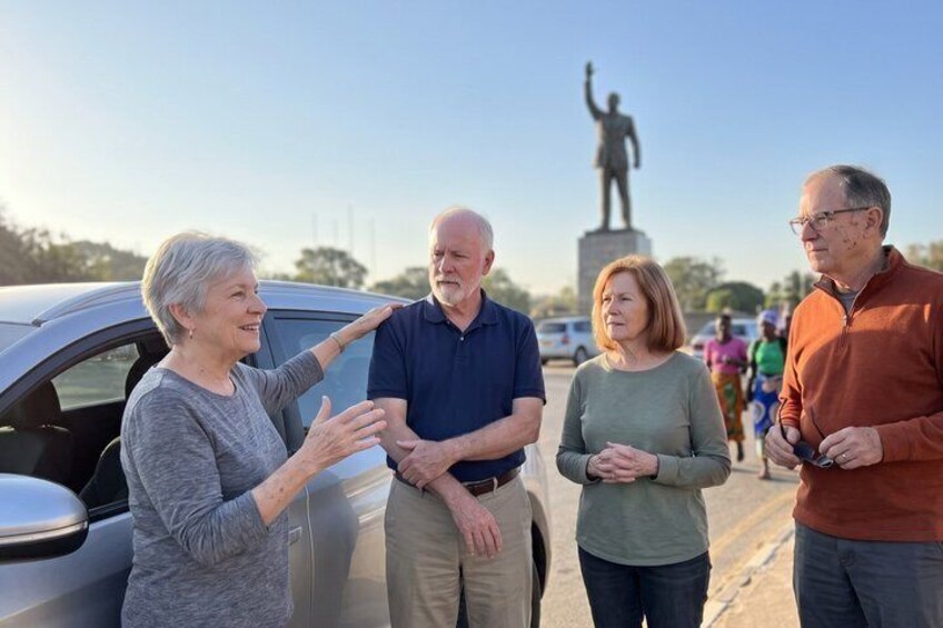 A group of people standing by a car and a statue in a park. 1