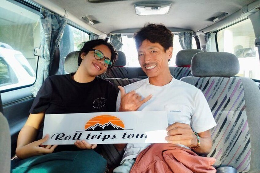 Two people sitting in a vehicle with a sign reading 'Roll trips tow' and a mountain graphic. 5