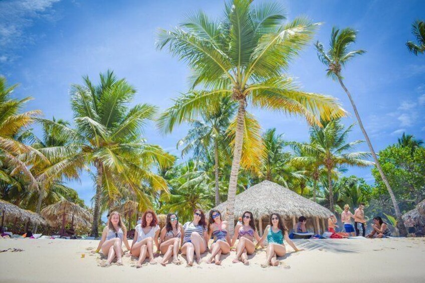 A group of people sitting on a beach with palm trees and a thatched hut in the background. 5