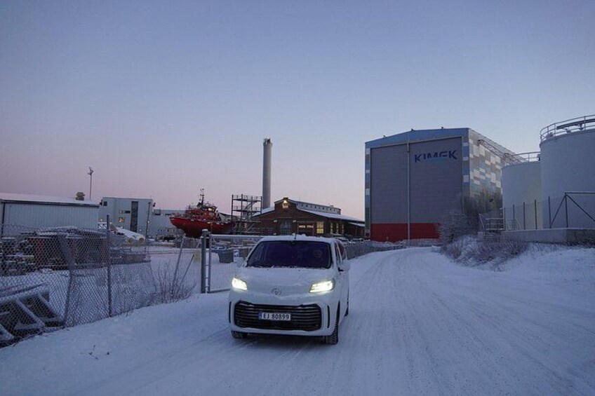 A white car driving on a snow-covered road in a snowy industrial area with buildings and a large chimney in the background. 5