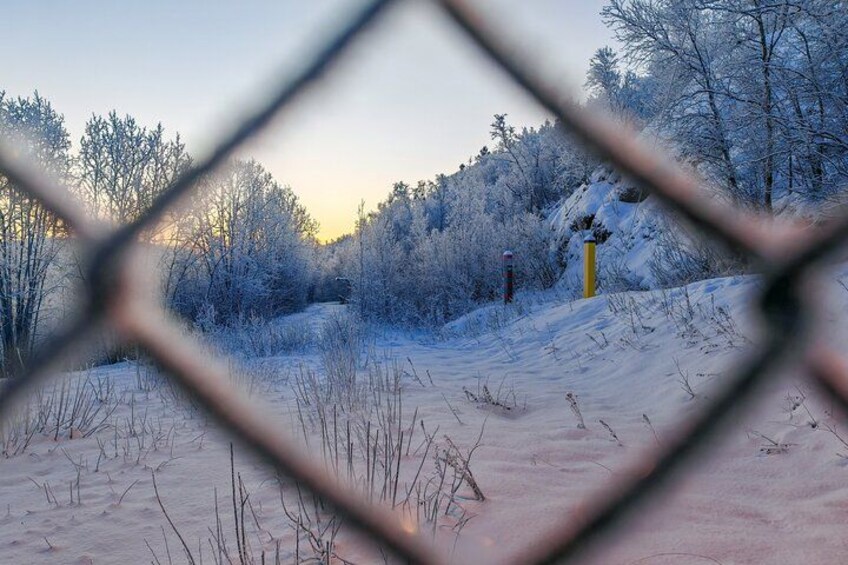 A snow-covered landscape with a fence in the foreground, a path leading through the trees, and a yellow pole in the distance. 3