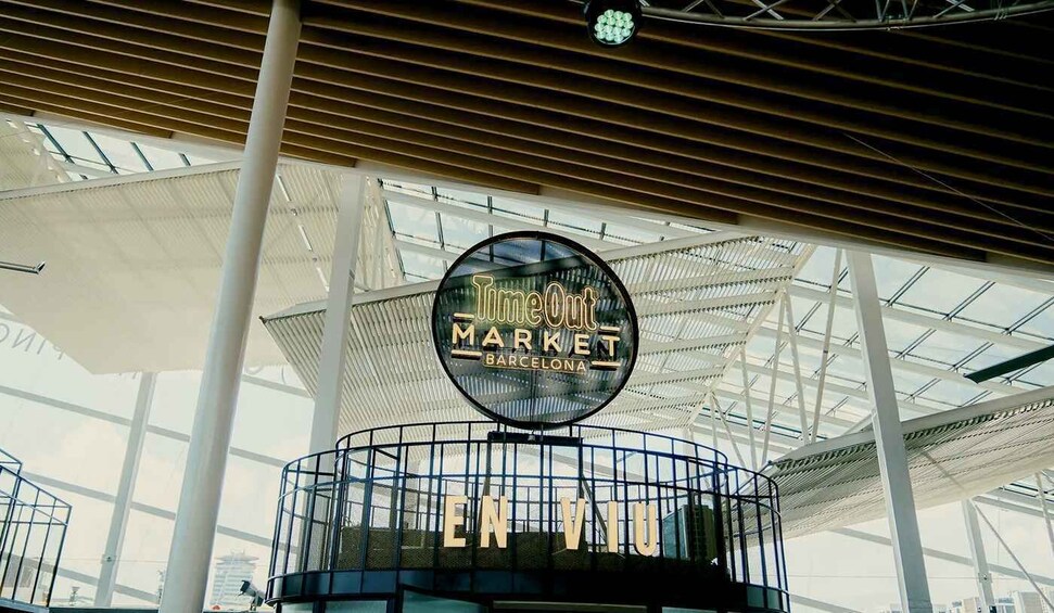 Een rond bord met 'Timeless Market Barcelona' erop, bevestigd aan een metalen constructie met een glazen overkapping. 4