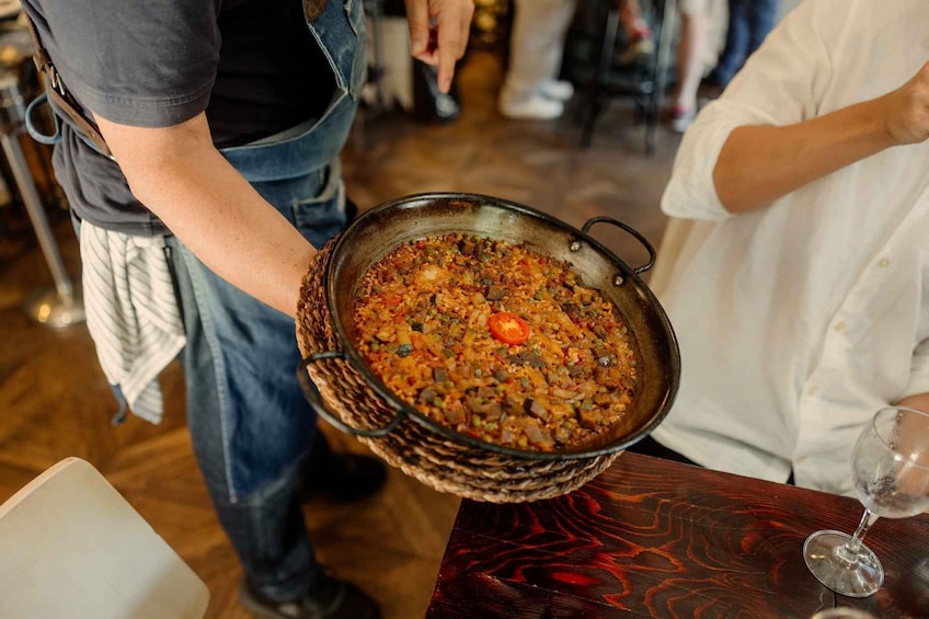 Een persoon houdt een grote pan eten vast, waarschijnlijk een stoofpot of ovenschotel, met een houten tafel en een wijnglas op de voorgrond. 1
