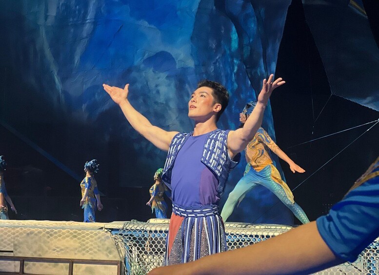 A person in a blue and purple costume with arms raised, standing on a stage with a backdrop of blue abstract shapes. 5