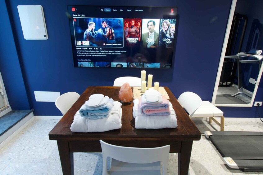 A room with a TV displaying movie posters, a table with towels, a salt lamp, and exercise equipment. 2